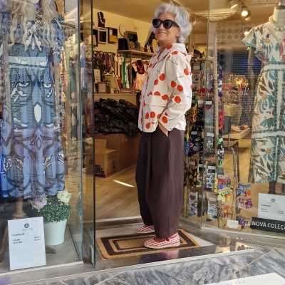 Senhora com camisa branca de bolinhas vermelhas e calças castanhas na entrada de loja de vestidos estampados