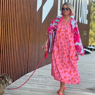 Mulher com vestido e casaco coloridos florais, bolsa listrada, a passear com cão em passadiço de madeira
