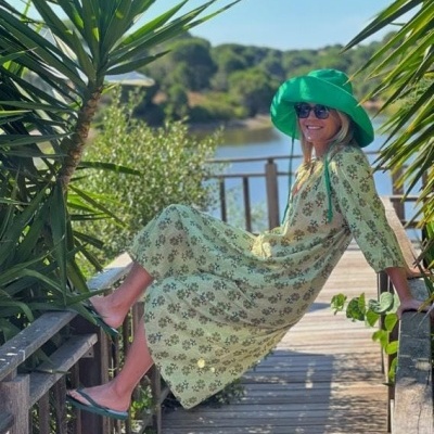 Mulher de vestido comprido floral verde e chapéu verde sentada numa passadeira de madeira ao ar livre