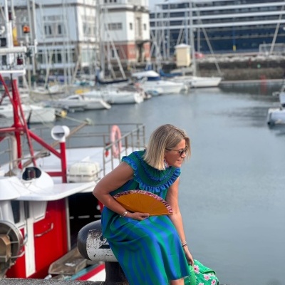 Mulher sentada no cais com vestido azul e verde, sapatos azuis e leque amarelo junto a barcos no porto.