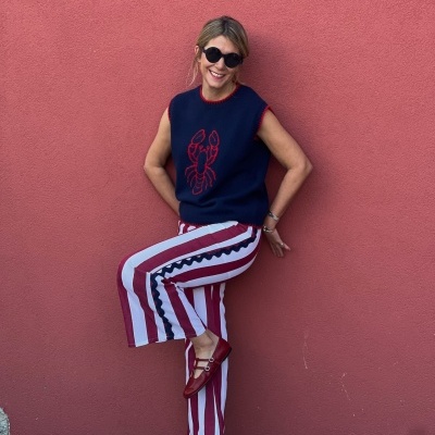 Mulher com camisola de malha azul escura com lagosta vermelha, calças às riscas e sapatos vermelhos, apoiada numa parede cor de rosa