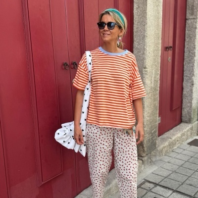 Mulher com t-shirt laranja e branco, calças brancas com padrão vermelho, sapatos laranja e mala branca com corações pretos junto a portas vermelhas.