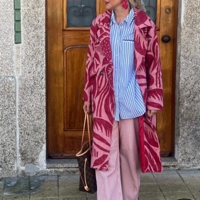 Mulher com casaco rosa estampado de folhas, camisa às riscas azul e branca, calças rosa, ténis vermelhos, bolsas e acessórios vermelhos à porta de madeira.
