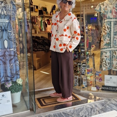 Mulher idosa com casaco branco de cerejas e calças castanhas em frente a loja de roupa