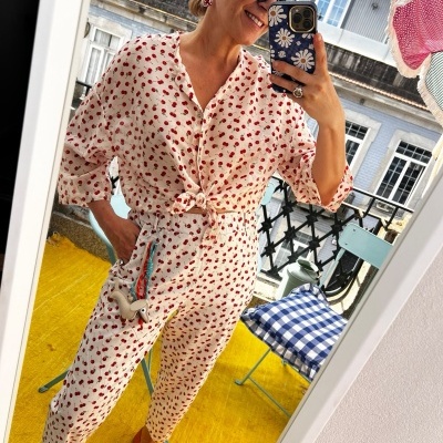 Conjunto branco com padrão de flores vermelhas, calças e túnica, sapatos laranja, fotografia em espelho com cadeira azul xadrez no fundo.