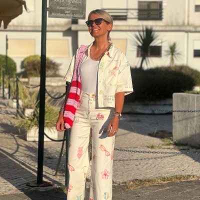 Mulher com roupa estampada bege claro, sandálias rosas, óculos escuros e bolsa vermelha e branca ao ar livre