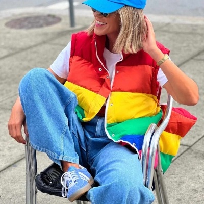 Mulher com colete colorido, calças de ganga, ténis brancos com azul e boné azul sentada em cadeira metálica ao ar livre