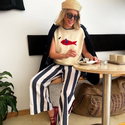 Mulher com roupa casual e chapéu sentada num café com calças às riscas e blusa com peixe vermelho.