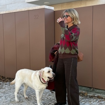 Mulher com camisola de padrão losangos e calças castanhas ao lado de um cão branco em frente a portão castanho número 32
