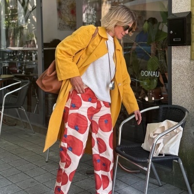 Mulher com casaco amarelo e calças floridas junto a cadeira preta com bolsa de tela branca.