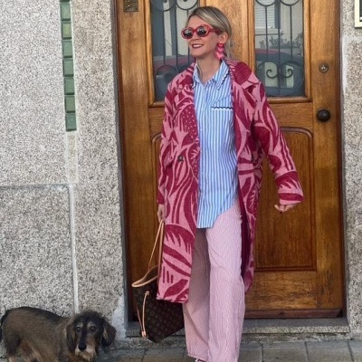 Mulher com casaco longo rosa e vermelho, camisa riscada azul clara, calças cor-de-rosa e sapatilhas vermelhas junto a uma porta de madeira e um cão pequeno