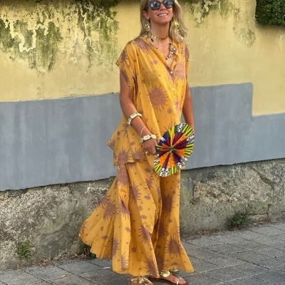 Mulher com conjunto amarelo mostarda e padrão castanho, sandálias douradas e leque colorido