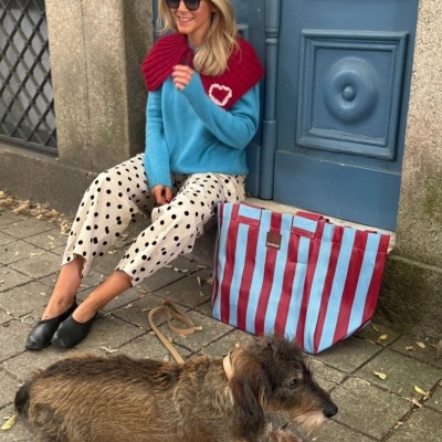 Mulher sentada com roupas coloridas e saco listrado ao lado de cão de pelo castanho em frente a porta azul