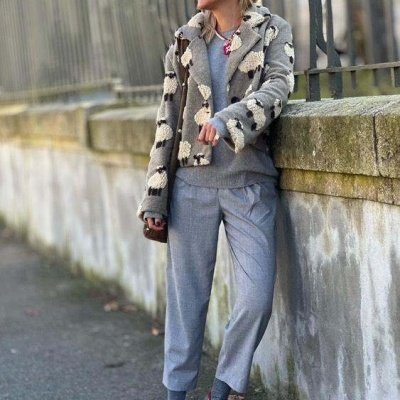 Mulher com casaco de padrão manchas cinza e branco, calças cinza e sapatos vermelhos junto a muro de pedra.