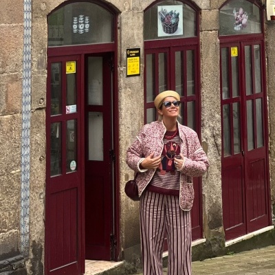 Mulher com casaco rosa floral e calças riscas junto a portas vermelhas num edifício azulejado