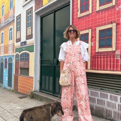 Pessoa com macacão rosa floral e camisa branca em frente a parede com mural colorido, ao lado de um cão.