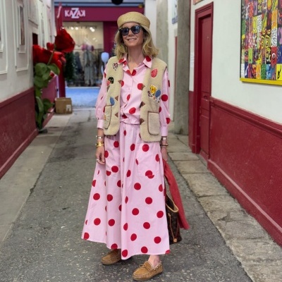 Mulher com roupa rosa de bolinhas vermelhas, colete bege, chapéu de palha e mocassins em rua com paredes vermelha e branca