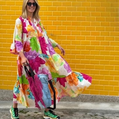 Mulher com vestido comprido floral colorido e ténis, parede amarela de azulejos atrás