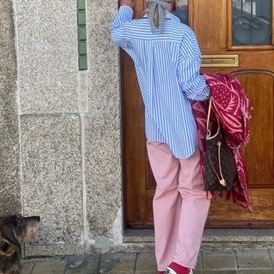 Mulher de costas vestindo camisa azul às riscas e calças cor-de-rosa em frente a porta de madeira, acompanhada por um cão