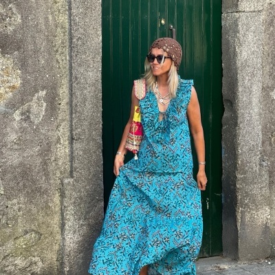 Vestido azul feminino com padrão floral, sem mangas e folhos, com sandálias castanhas e chapéu castanho, em frente a porta verde