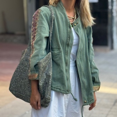 Pessoa com casaco verde com padrões vermelhos e bege, vestido branco e mala de tecido verde com padrão bege