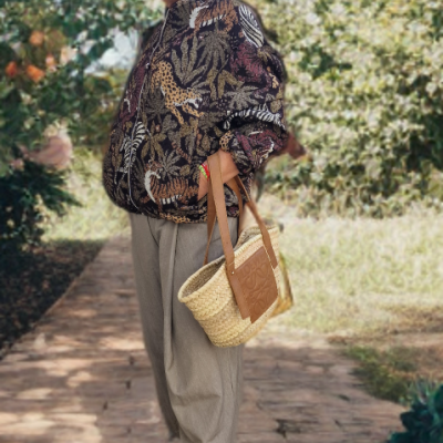 Senhora idosa com casaco estampado, calças cinza, sandálias castanhas e bolsa de palha ao ar livre