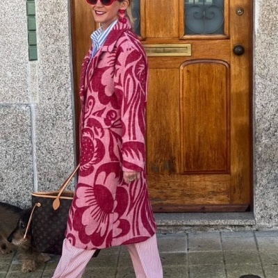 Mulher de casaco rosa com padrão floral vermelho e ténis vermelhos junto a uma porta de madeira