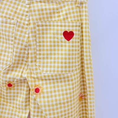 Calças amarelo e branco vichy com bordados de coração e flores penduradas num cabide.