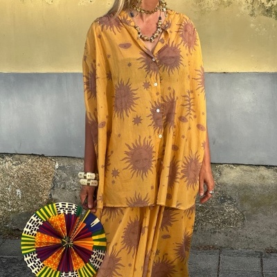 Conjunto amarelo com padrão de sóis, mulher com leque colorido de mão em ambiente urbano.