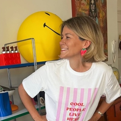 Mulher com t-shirt branca com texto vermelho, brincos de morangos, estante com objetos coloridos e smiley amarelo ao fundo.