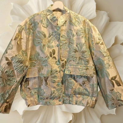 Casaco jacquard dourado e verde com padrão floral, botão frontal e bolsos, pendurado num cabide de madeira