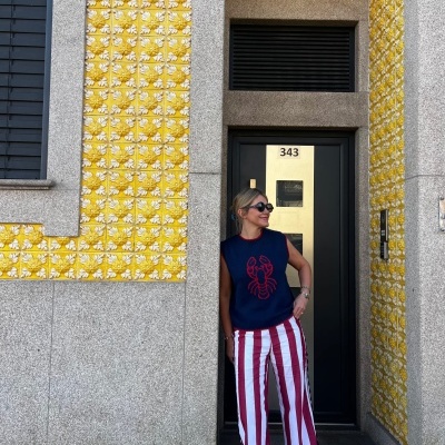 Mulher com colete azul e calças às riscas vermelhas e brancas numa porta com azulejos amarelos