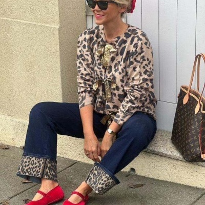 Mulher sentada com casaco animal print, calças de ganga, sapatilhas vermelhas e mala castanha junto a porta branca.