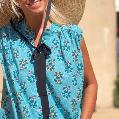 Blusa azul-turquesa com padrão floral e botões brancos usada por mulher com chapéu de palha e óculos de sol