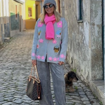 Mulher com casaco bordado e cachecol rosa numa rua de pedra, segurando mala e com cão ao lado