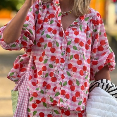 Blusa feminina xadrez rosa com cerejas vermelhas e calças iguais, segurando casaco branco acolchoado.