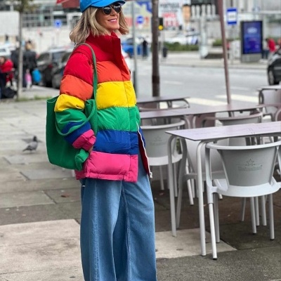 Pessoa com casaco acolchoado arco-íris e calças de ganga na rua junto a esplanada com chapéus de sol vermelhos
