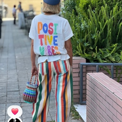 Pessoa com t-shirt branca escrita POSITIVE VIBES, calças riscas coloridas e mala de fio trançado colorido.