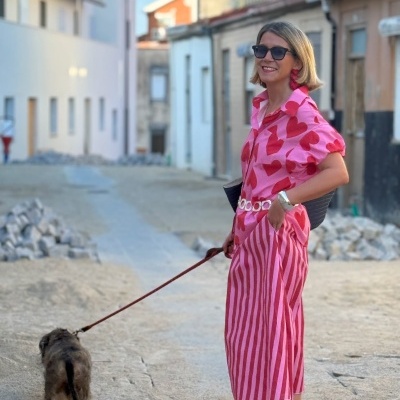 Mulher de vestido rosa com corações vermelhos e riscas, óculos de sol e sandálias vermelhas a passear com cão numa rua.