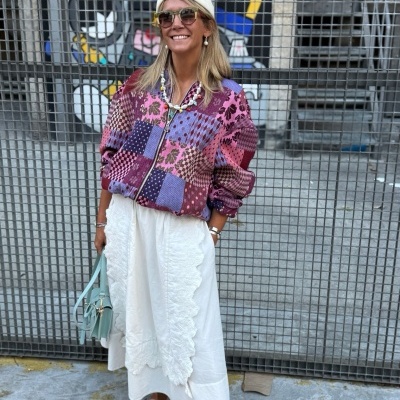 Mulher com casaco patchwork colorido, saia branca rendada e chapéu branco, segurando mala azul clara com fundo de grade metálica e grafite