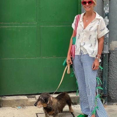 Mulher com camisa branca estampada, calças xadrez com fitas verdes e óculos vermelhos, a passear cão pequeno.