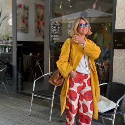Mulher loira com casaco amarelo e calças largas estampadas posa em café ao ar livre.