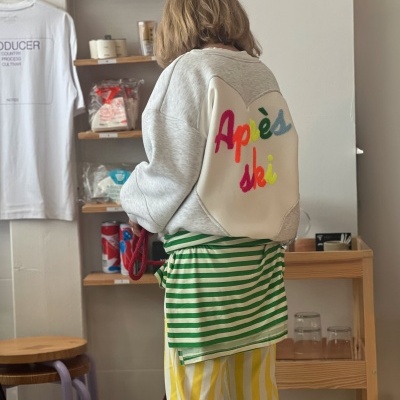 Pessoa de costas com camisola cinzenta com texto colorido atrás e calças riscas amarelas e brancas em interior com prateleiras e t-shirt pendurada