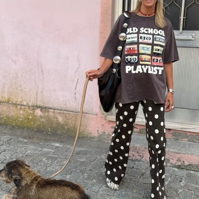 Mulher com t-shirt 'OLD SCHOOL PLAYLIST', calças de bolinhas, lenço vermelho e óculos a segurar trela de cão castanho na rua