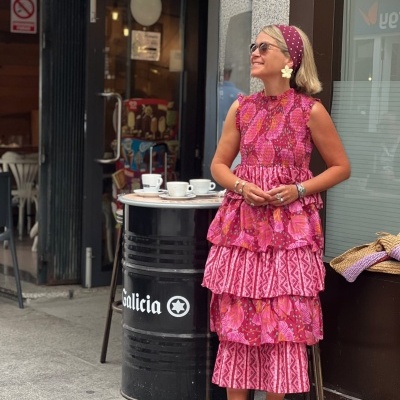Mulher com vestido floral rosa e vermelho e acessório de cabelo vermelho de bolinhas junto a barril com mesa e chávenas