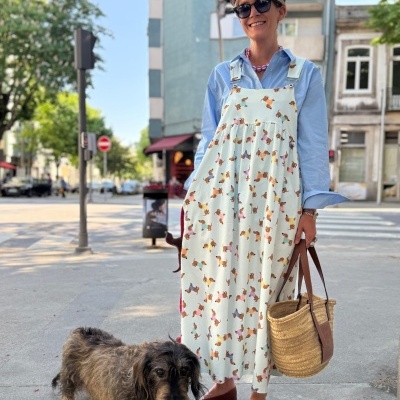 Pessoa com vestido branco com padrão de cães coloridos, chapéu castanho e óculos escuros, segurando bolsa de palha, com cão ao lado