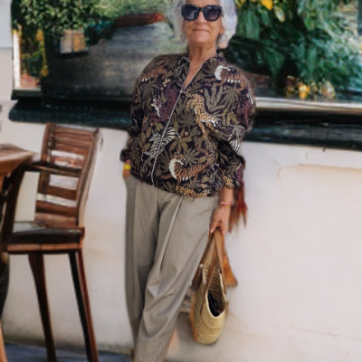 Mulher com casaco estampado, calças bege e mala de palha em espaço exterior com flores.