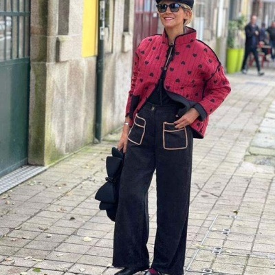 Mulher com casaco vermelho com padrão de coroas, calças pretas largas e boina preta numa rua citadina.