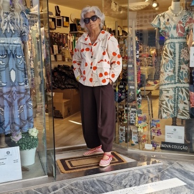 Mulher de casaco branco com bolas vermelhas e calças castanhas à porta de loja com manequins e texto NOVA COLEÇÃO