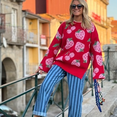 Mulher com camisola vermelha de morangos e calças azuis riscas posando numa rua antiga